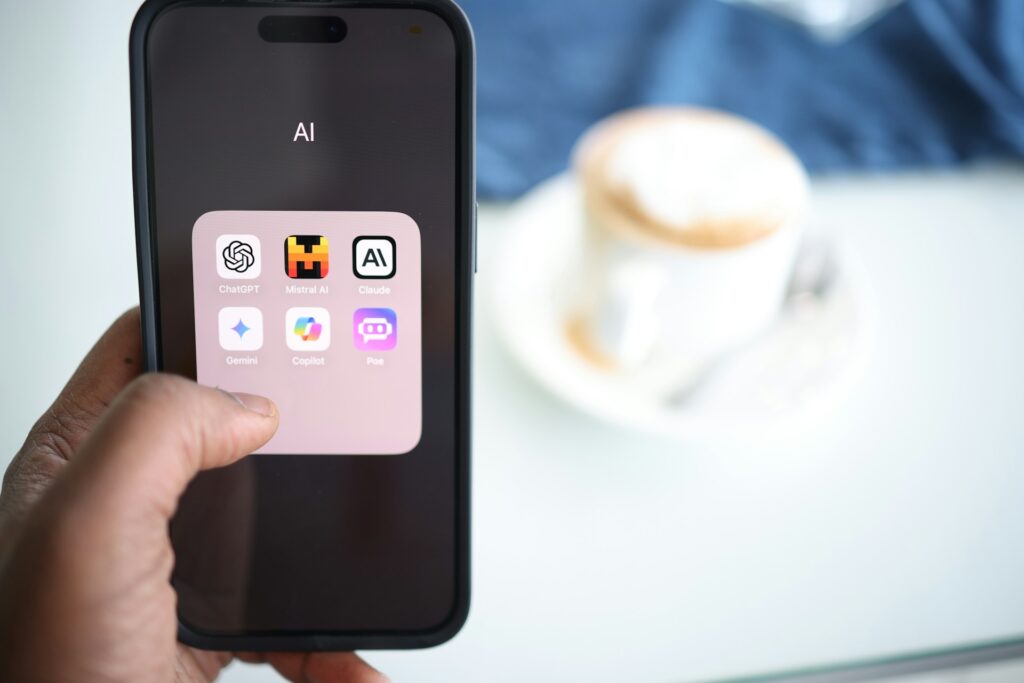 Close-up of an iPhone screen showing an AI apps folder with ChatGPT, Mistral AI, Claude, Gemini, Copilot, and Poe; blurred background coffee cup nearby.