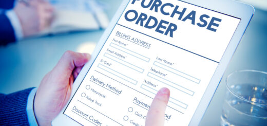 Tablet screen displaying a purchase order form with billing/shipping fields and payment options, being filled by a hand on a blue-toned desk surface.