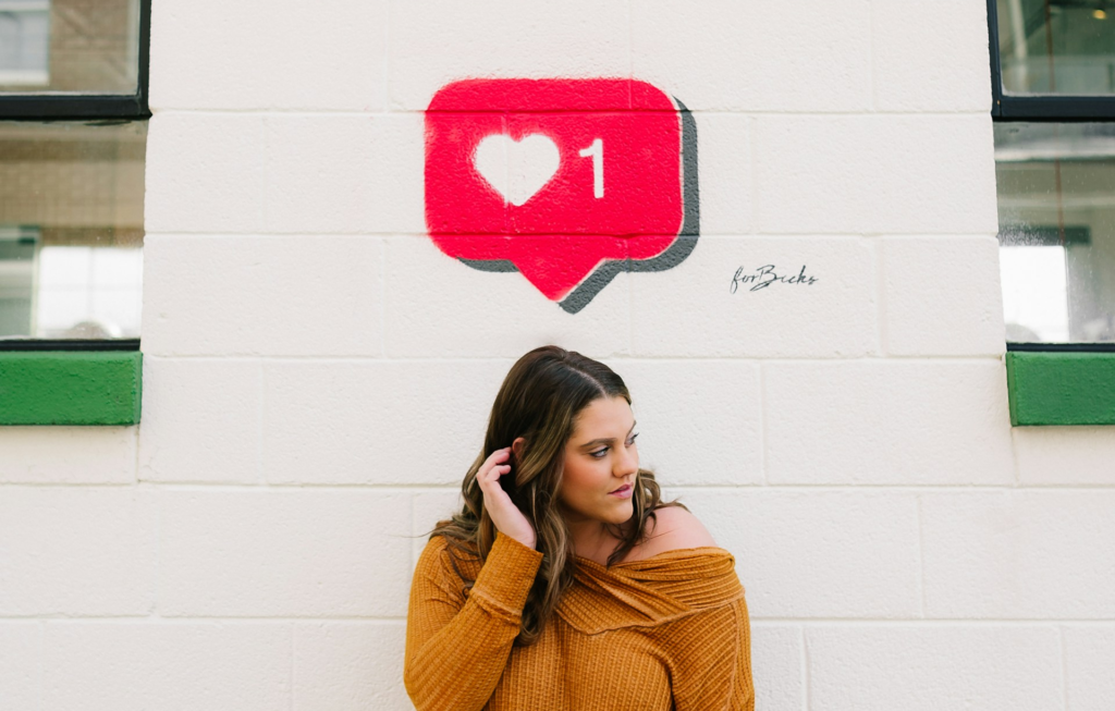 The Algorithm Stopped Counting Likes 2 - SaltMinePublishing.com Woman in an orange knit sweater stands against a white wall with a graffiti-style pink heart notification bubble showing the number 1 above her head.
