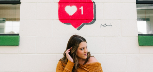 Woman in an orange knit sweater stands against a white wall with a graffiti-style pink heart notification bubble showing the number 1 above her head.