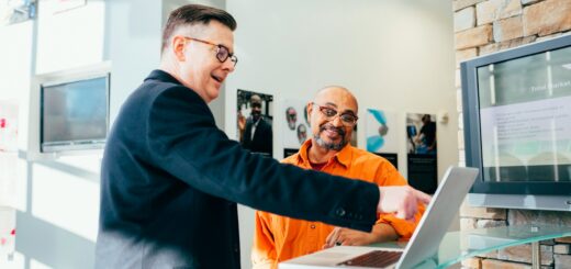 Two men discuss at a showroom desk, one in a dark suit pointing to a laptop screen beside a brick feature wall and display monitor behind them